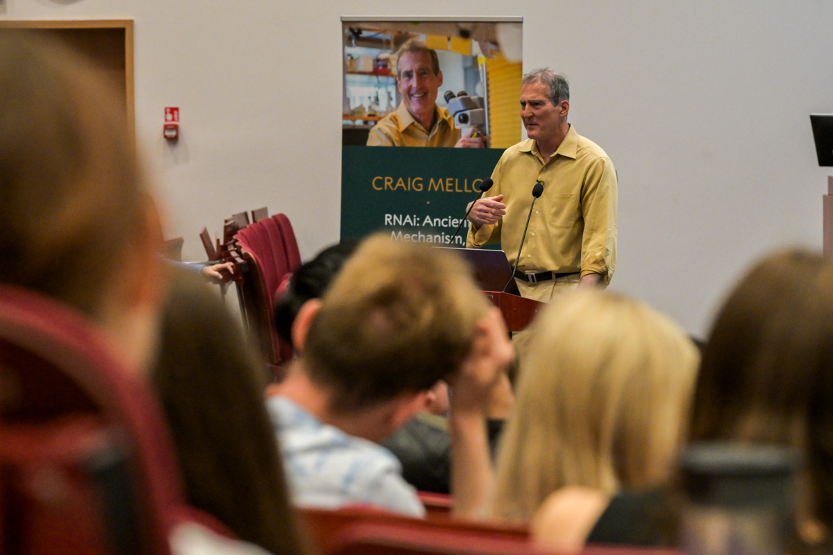 Prof. Craig C. Mello, man in a yellow shirt  with a microphone stands in front of an audience in a lecture hall. He gestures with his hand. A poster in the background reads 'CRAIG MELLO RNA: Ancient Mechanisms.