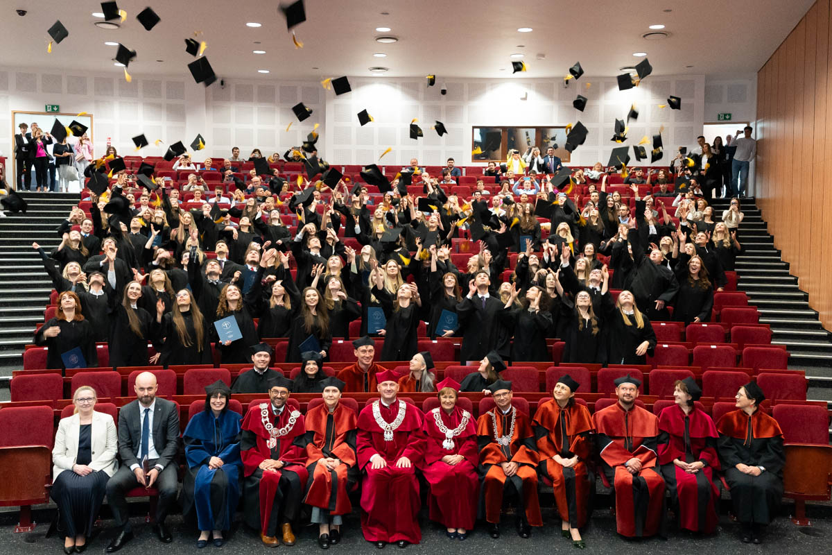 The graduation ceremony takes place in a large, brightly lit auditorium. In the center is a group of graduates dressed in traditional black togas and birettas, who cheerfully toss their caps up in the air. The graduates stand in several rows of red armchairs, with other ceremony participants visible behind them. In the foreground, in front of the rows of graduates, sits a group of people in formal academic attire.