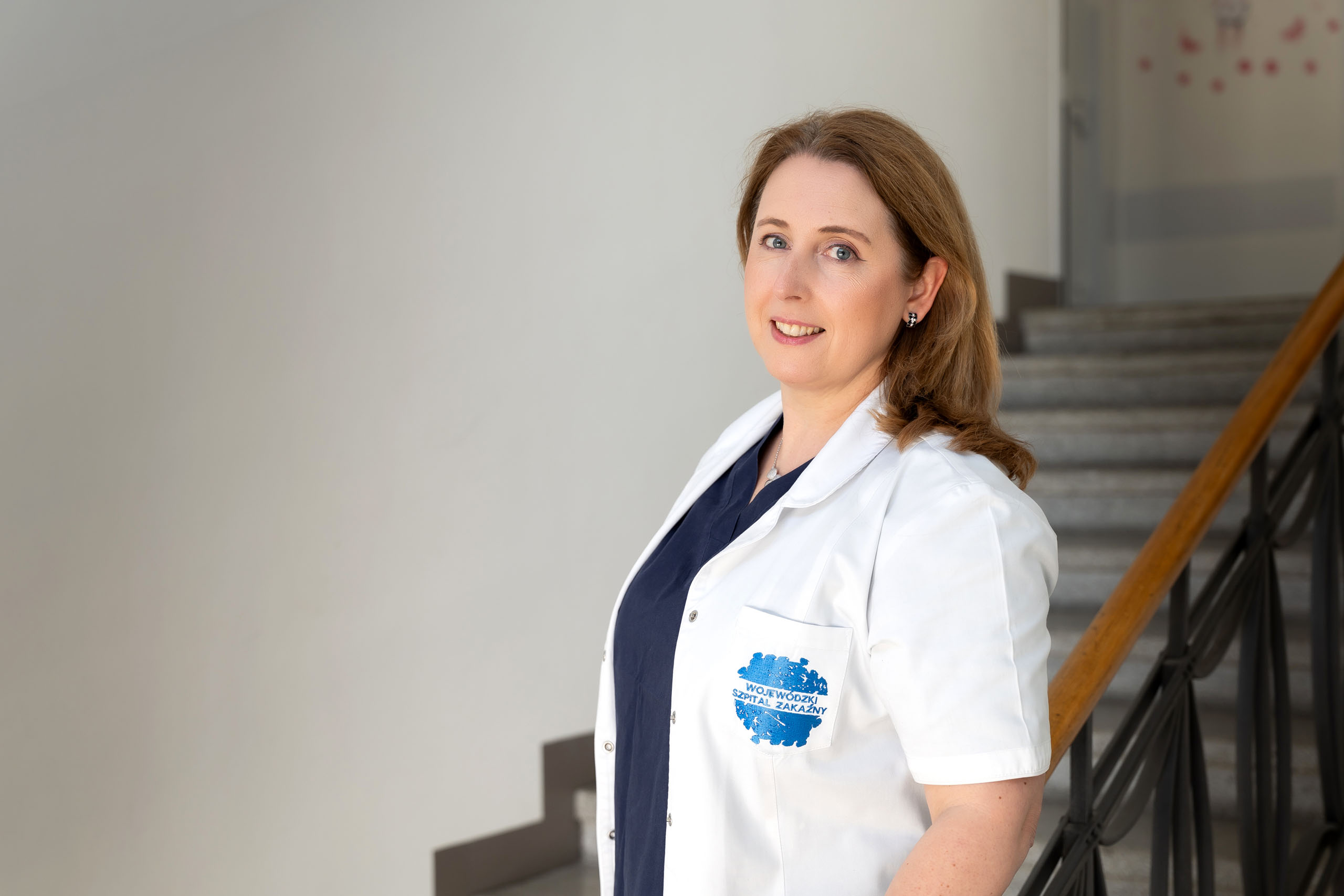The woman is standing inside the building next to the stairs. She is wearing a white lab coat with an emblem on the left side that includes text and a blue circular design. The background of the photo shows part of the staircase with metal handrails and steps leading up.