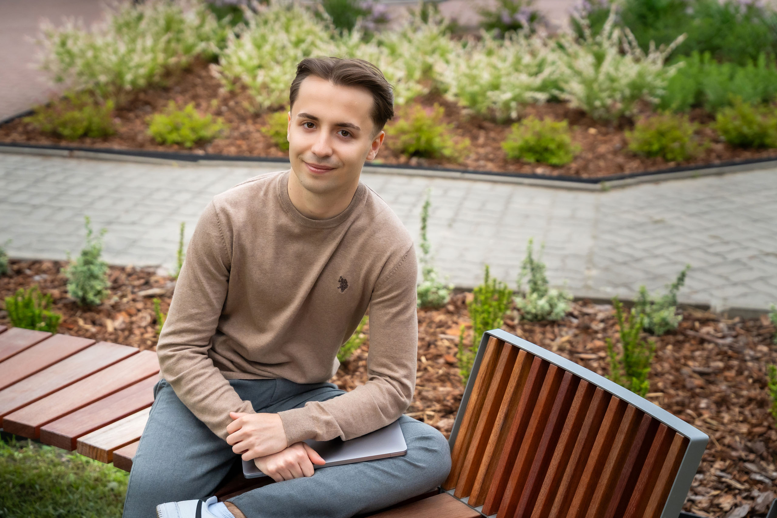 A young man, Adrian Bednarek, looks into the camera lens and smiles for the photo. He has dark hair and dark eyes. He is in a brown sweater and gray pants sitting on a wooden park bench, holding a laptop on his lap. Visible in the background is a neat flower bed with plants and a cobblestone sidewalk.