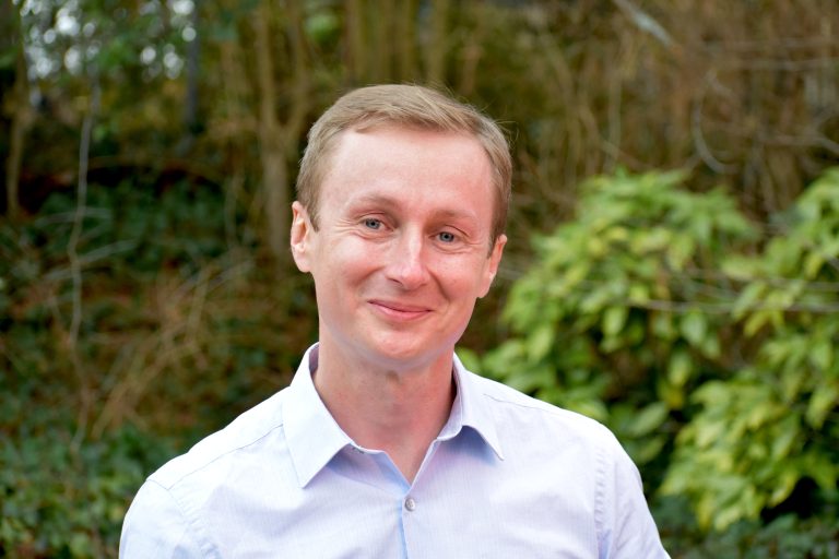 Mężczyzna prof. Mazurowski w jasnoniebieskiej koszuli, z jasnymi włosami uśmiecha się do zdjęcia. Stoi na zewnątrz. W tle widać zielone krzewy i drzewa.