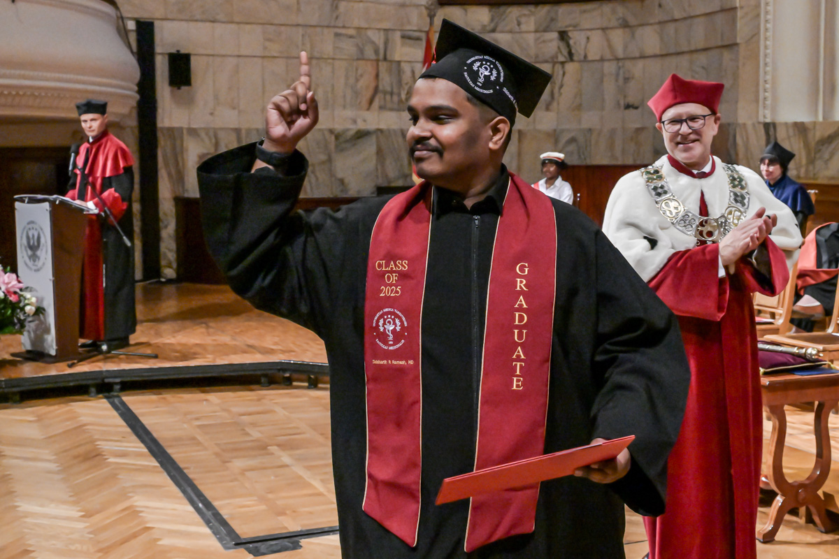 The graduate, wearing a black toga and biretta with a red scarf inscribed with ‘CLASS OF 2023’ and ‘GRADUATE’, holds his diploma in his left hand. He raises his right hand with his index finger extended. In the background people in togas, one is speaking at the rostrum.
