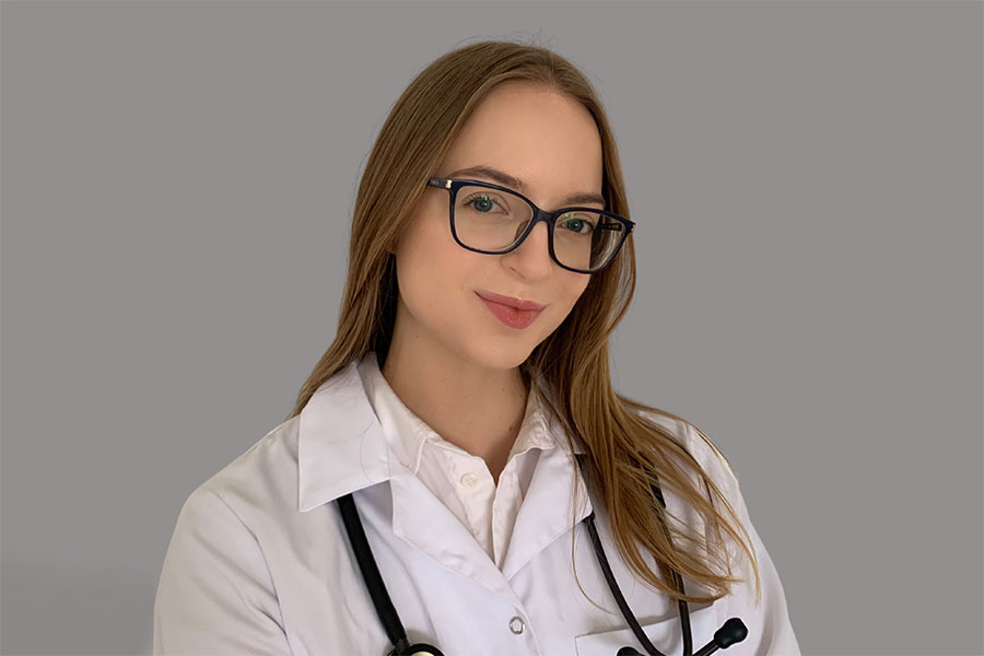 A young woman wearing glasses and a white doctor's apron with a stethoscope around her neck, against a grey background. She smiles at the photograph.