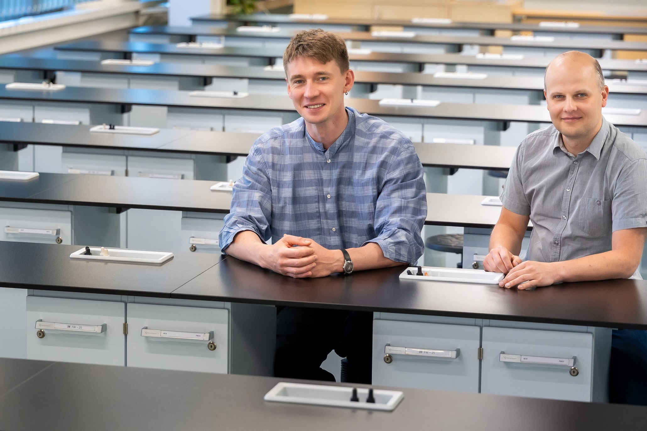 Two men: from the left Professor Piwowarski, from the right Professor Granica,sit in a bright, modern training room. The interior features rows of desks with black tops and white drawers.