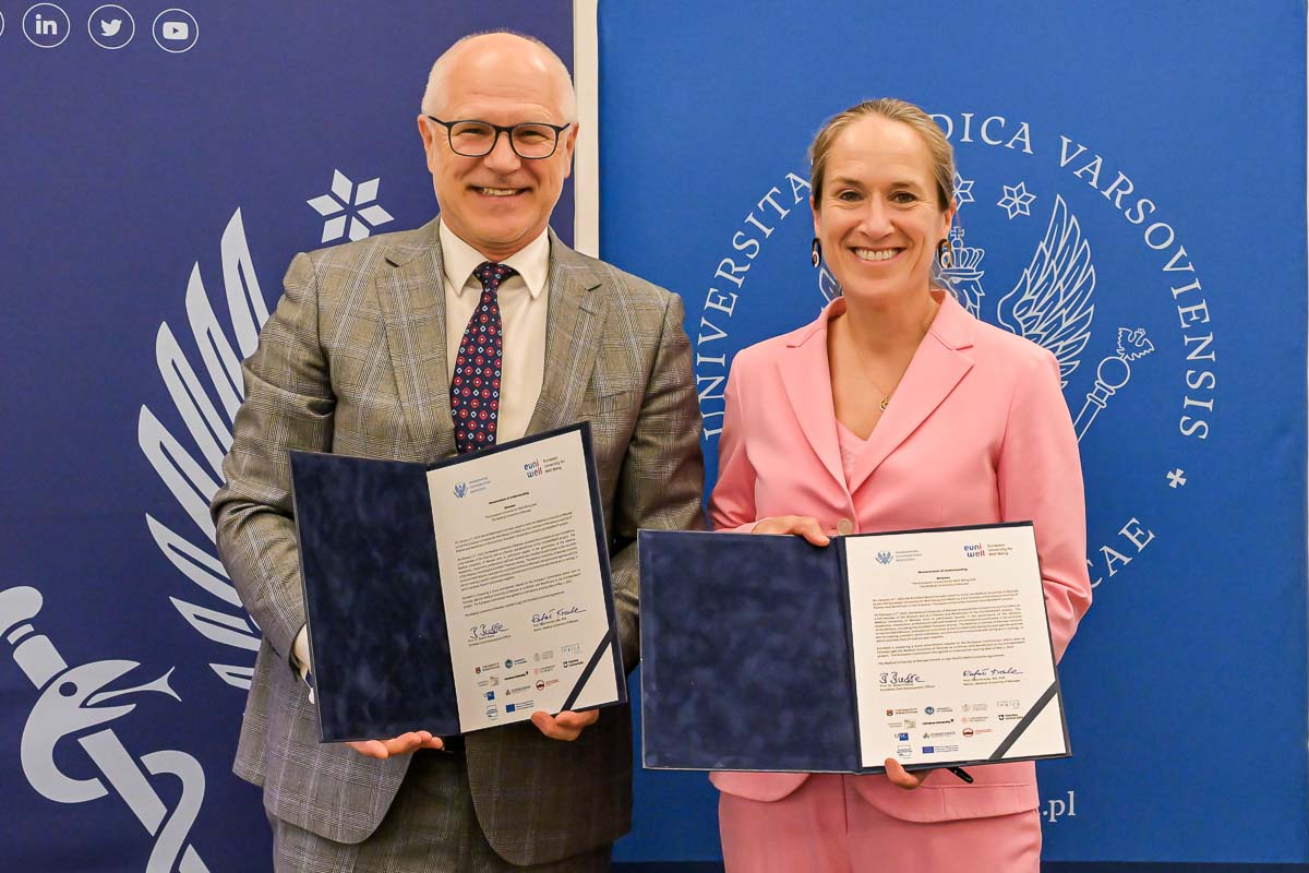 A man in a grey suit and a woman in a pink suit are presenting documents in open folders. Two banners with the logo of the Medical University of Warsaw are visible in the background.