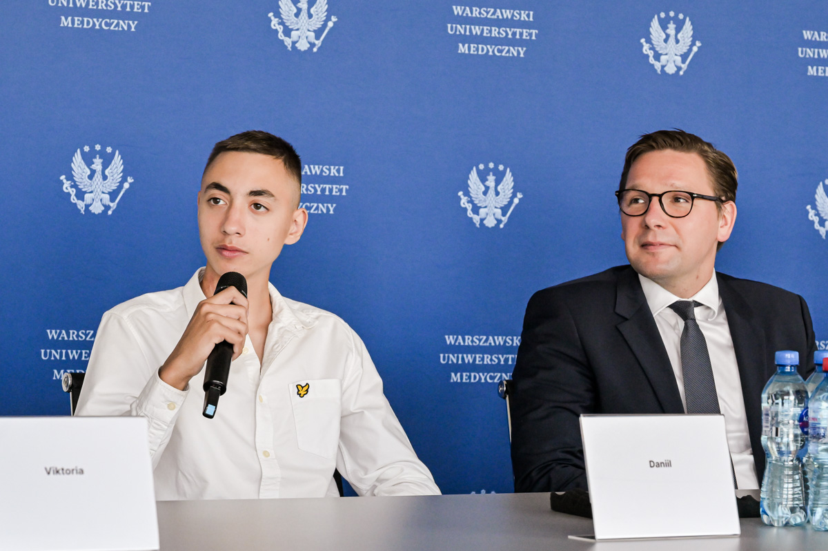 On the right, a young man in a white shirt with an embroidered logo on the chest is sitting and holding a microphone. Next to him is a man in a dark suit, white shirt, and tie. Both are sitting at a table during a press conference.