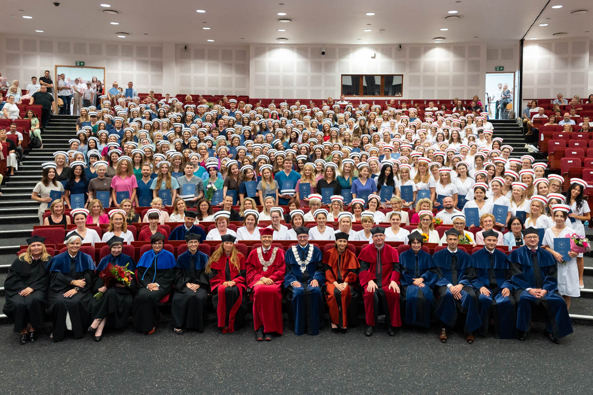 The capping ceremony for nursing and midwifery graduates in the auditorium. In the front row sit people in academic gowns—the teaching staff. Behind them stand the graduates in colorful outfits, wearing nursing and midwifery caps and holding their diplomas.