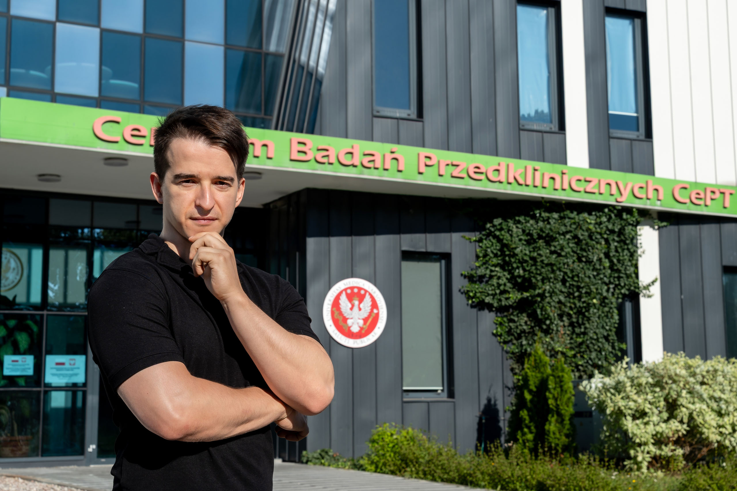 Mateusz Rybka, a man wearing a black T-shirt, is standing in front of the CePT Preclinical Research Center building. He is resting his chin on his left hand. He is looking at the photo. The building has gray walls and large windows. Above the entrance is a green sign with the name. Next to the door is a red coat of arms with a white eagle. In the background are decorative plants.