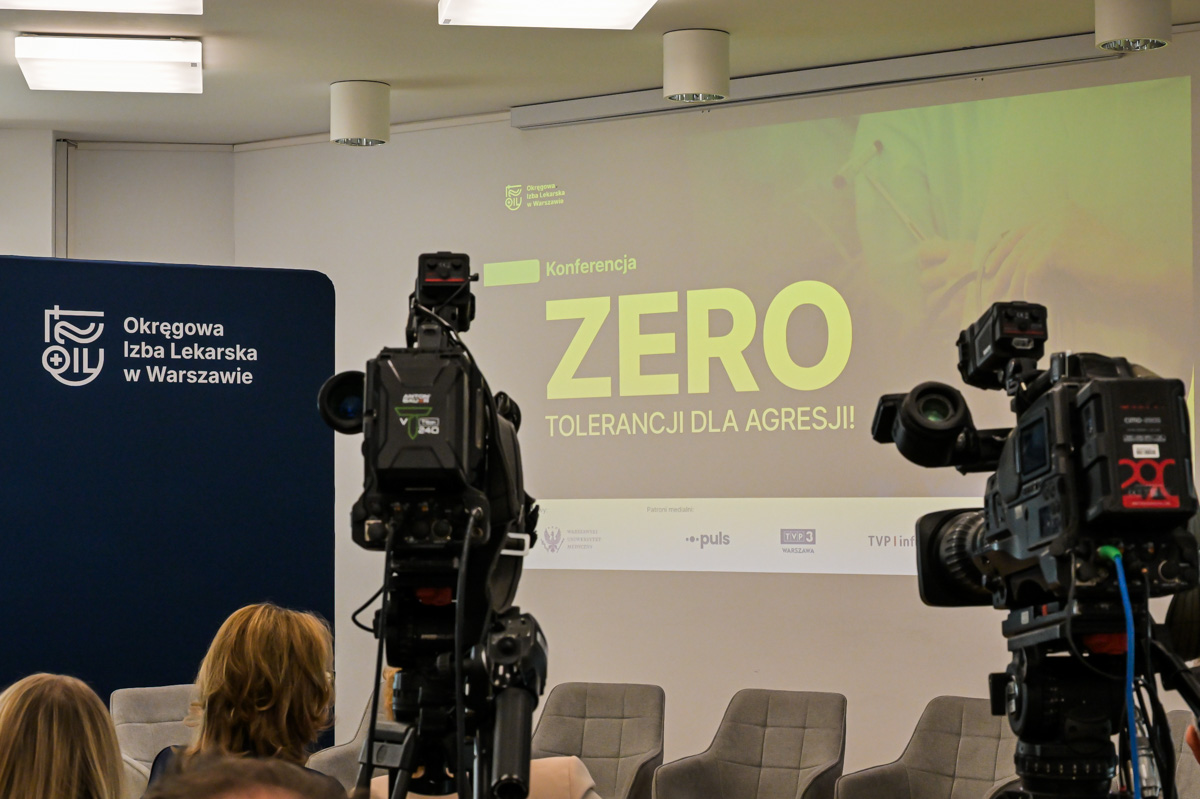A screen with the words “zero tolerance for aggression” and two cameras. Next to the screen, there is a blue wall with the white logo of the Regional Medical Chamber. 