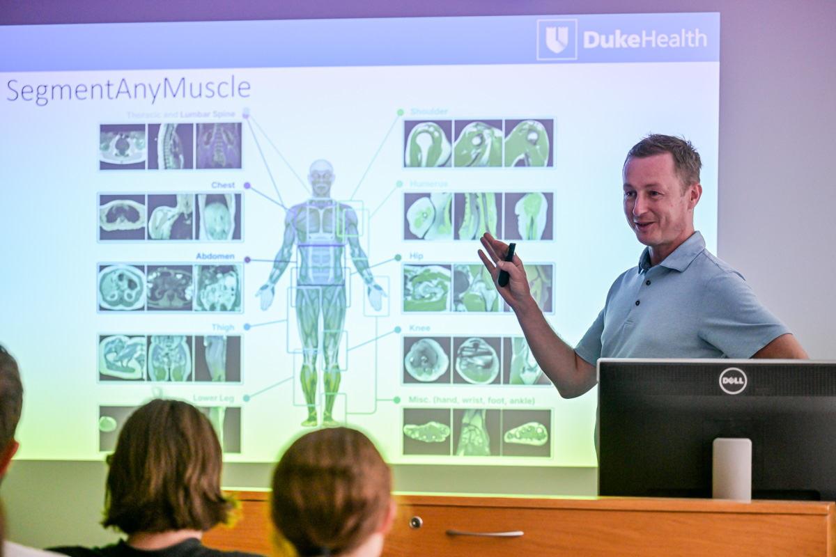 Medical presentation in a lecture hall. On the screen, a slide titled “SegmentAnyMuscle” and the DukeHealth logo. A colorful illustration of human muscles and MRI images of the neck, chest, abdomen, thigh, knee, and foot are visible. The presenter points to the slide. In the background, audience members are seated, visible from behind.