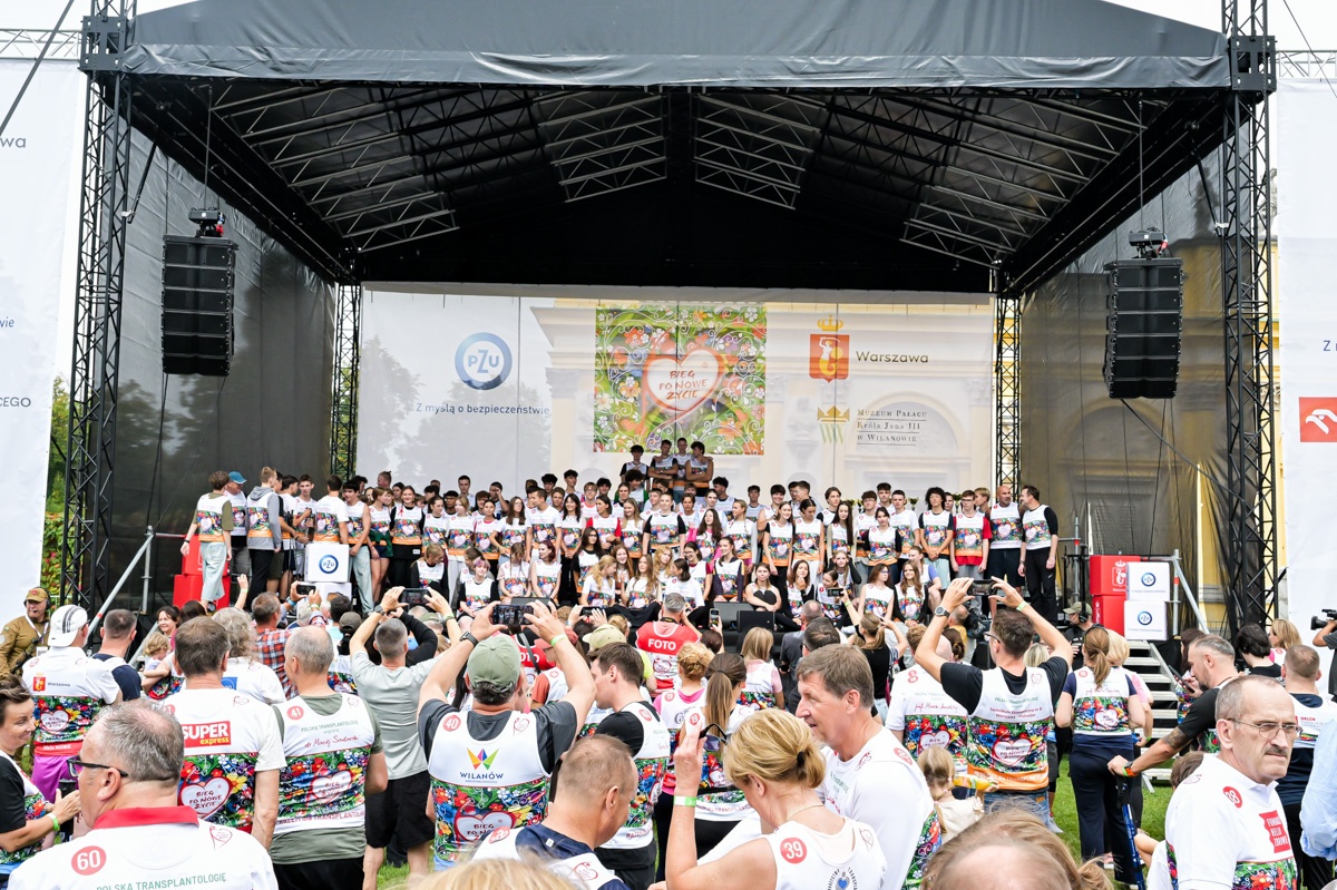 There are about 30 people on stage, wearing sportswear and t-shirts with start numbers. There is a crowd of people in front of the stage, some of them taking photos.
