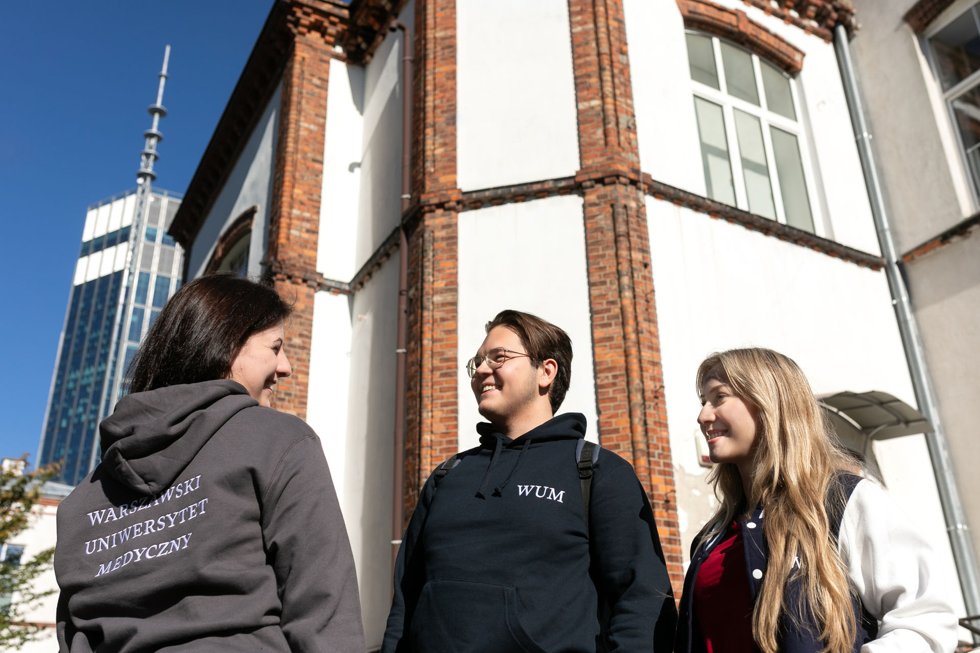 Three people are standing in front of a building. The person on the left is wearing a gray sweatshirt with the words “Warsaw Medical University” on it. A building can be seen in the background.