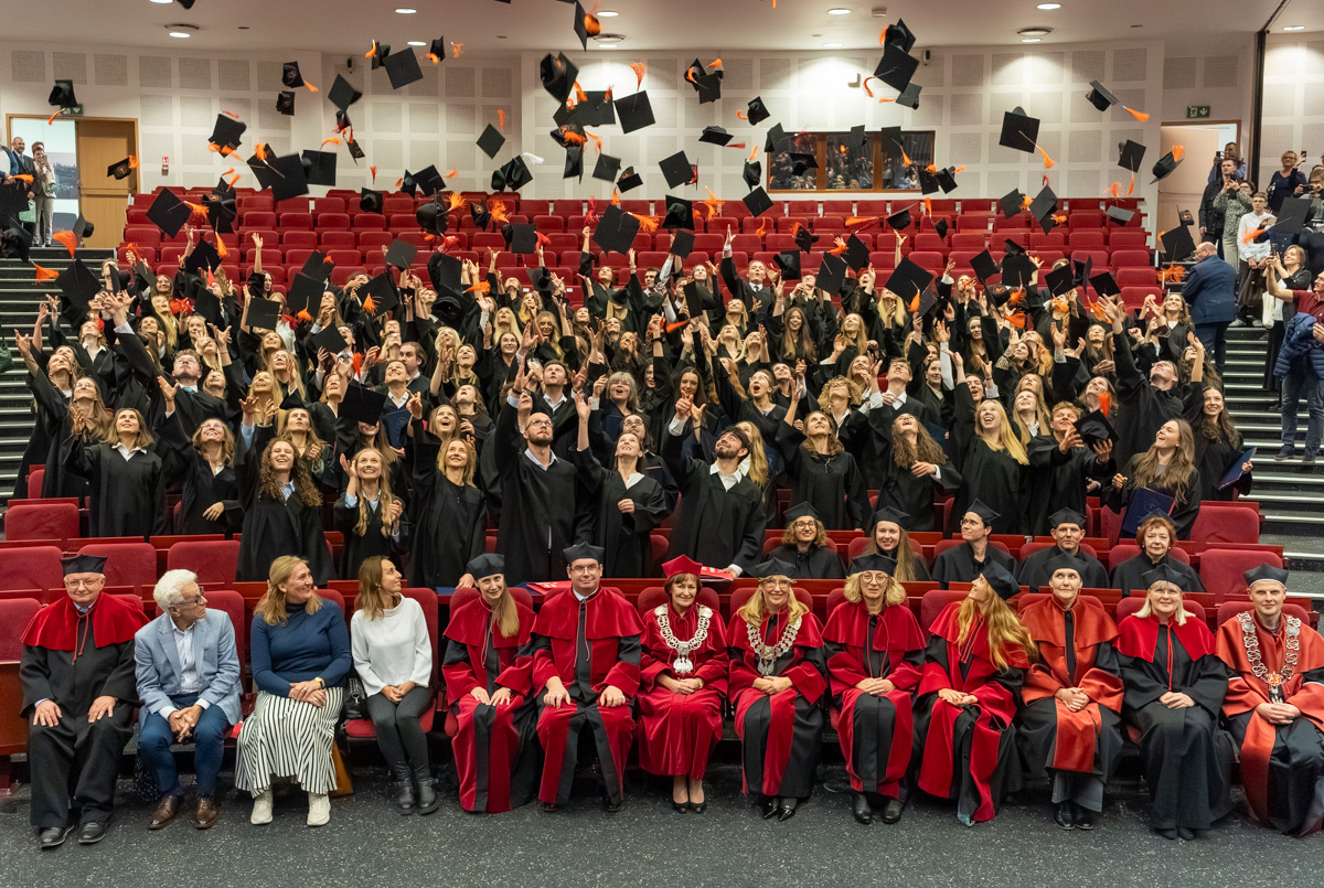 Graduates in black gowns throw their caps into the air; in the foreground, people in red gowns.