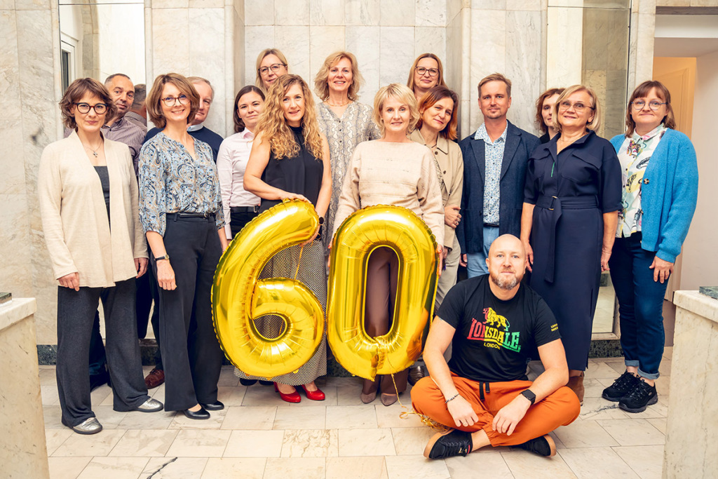 Grupa kobiet i mężczyzn stoi w jasnym pomieszczeniu z marmurowymi ścianami. W centrum dwie kobiety trzymają duże, złote balony w kształcie liczby „60”.