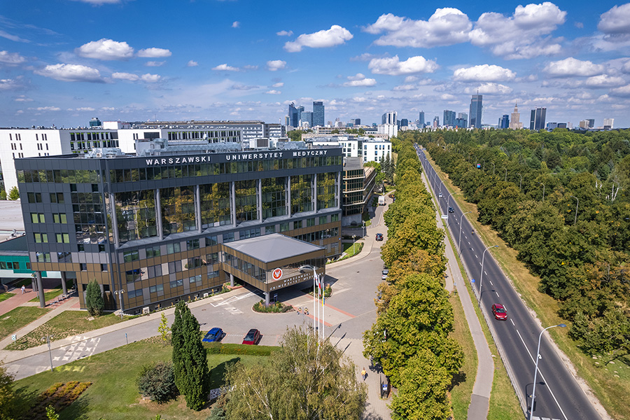 In the foreground, there is a modern building with large windows and the words “Warsaw Medical University” on the roof. There are flags in front of the building. On the right, there is a wide street surrounded by trees. In the background, there is a panorama of the city with skyscrapers and office buildings.