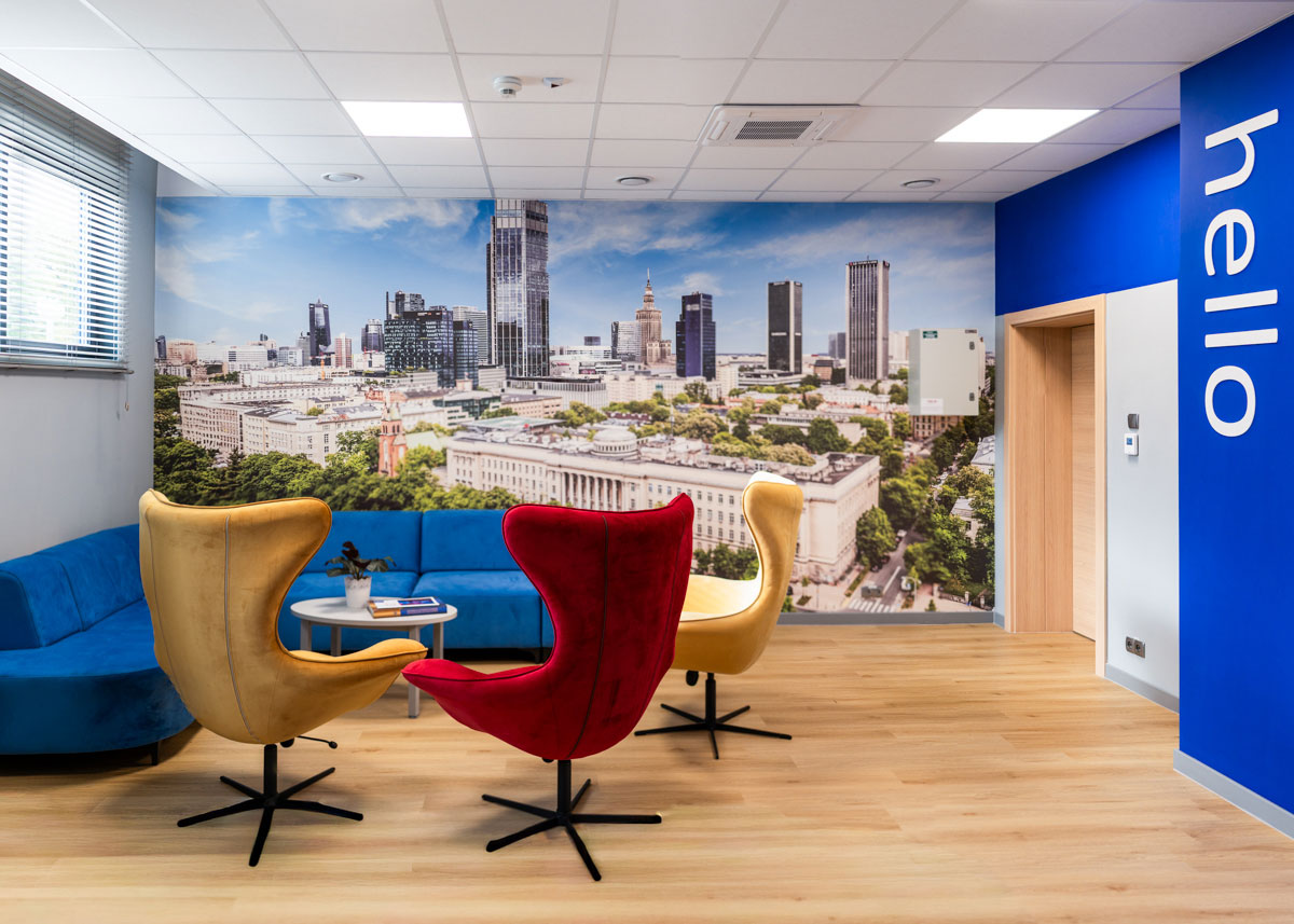 A bright office room with wooden parquet flooring and a large photo wallpaper of a city with skyscrapers. Four armchairs—two yellow, one red, and one blue—are arranged around a round table. On the left side, there is a blue sofa. On the right wall, there is a “hello” sign on a blue background. Daylight streams in through a large window with blinds.