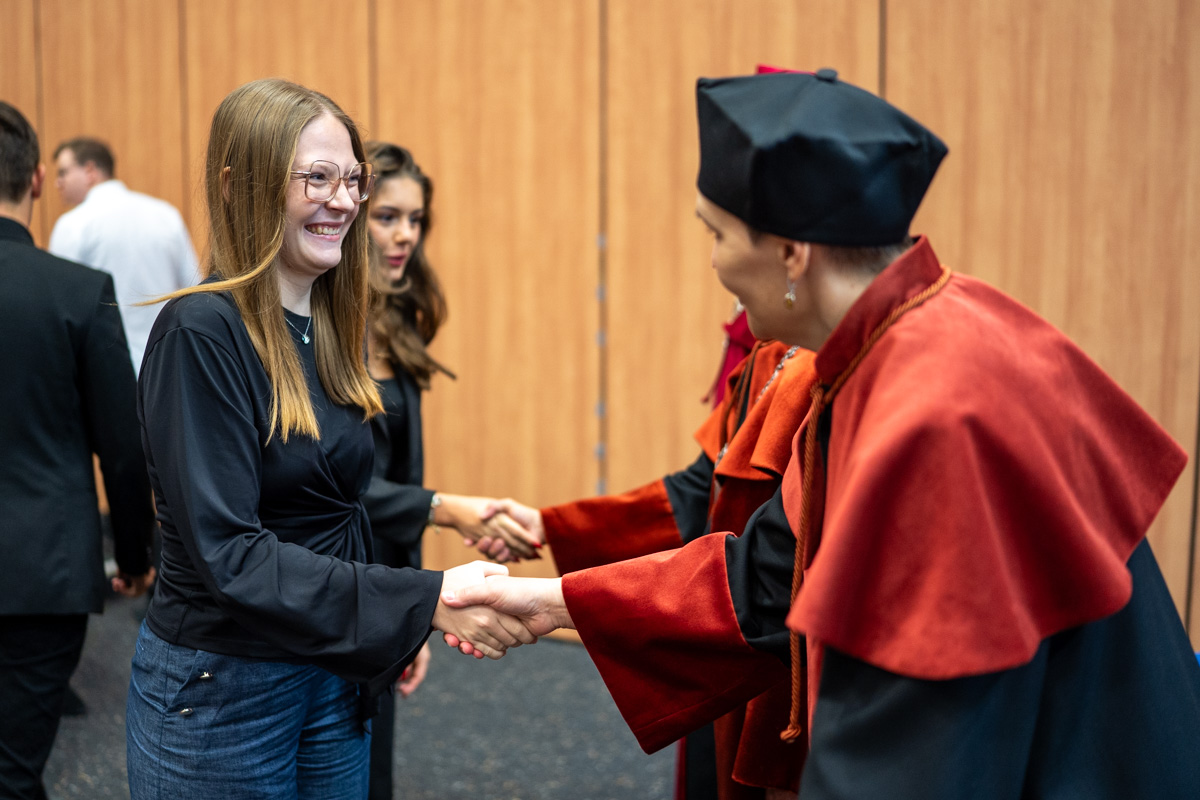 The vice dean for teaching in the field of pharmacy, dressed in a black and orange gown, shakes hands with a young girl—a student wearing a black blouse and jeans. The student smiles broadly. Two people in gowns stand next to them. Other people can also be seen in the background.