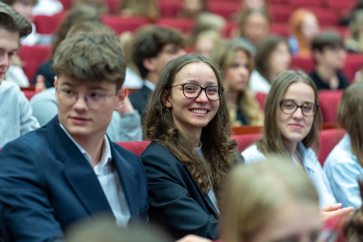 A group of young people sitting on red armchairs. Among them, two young girls look straight into the camera and smile for the photo.
