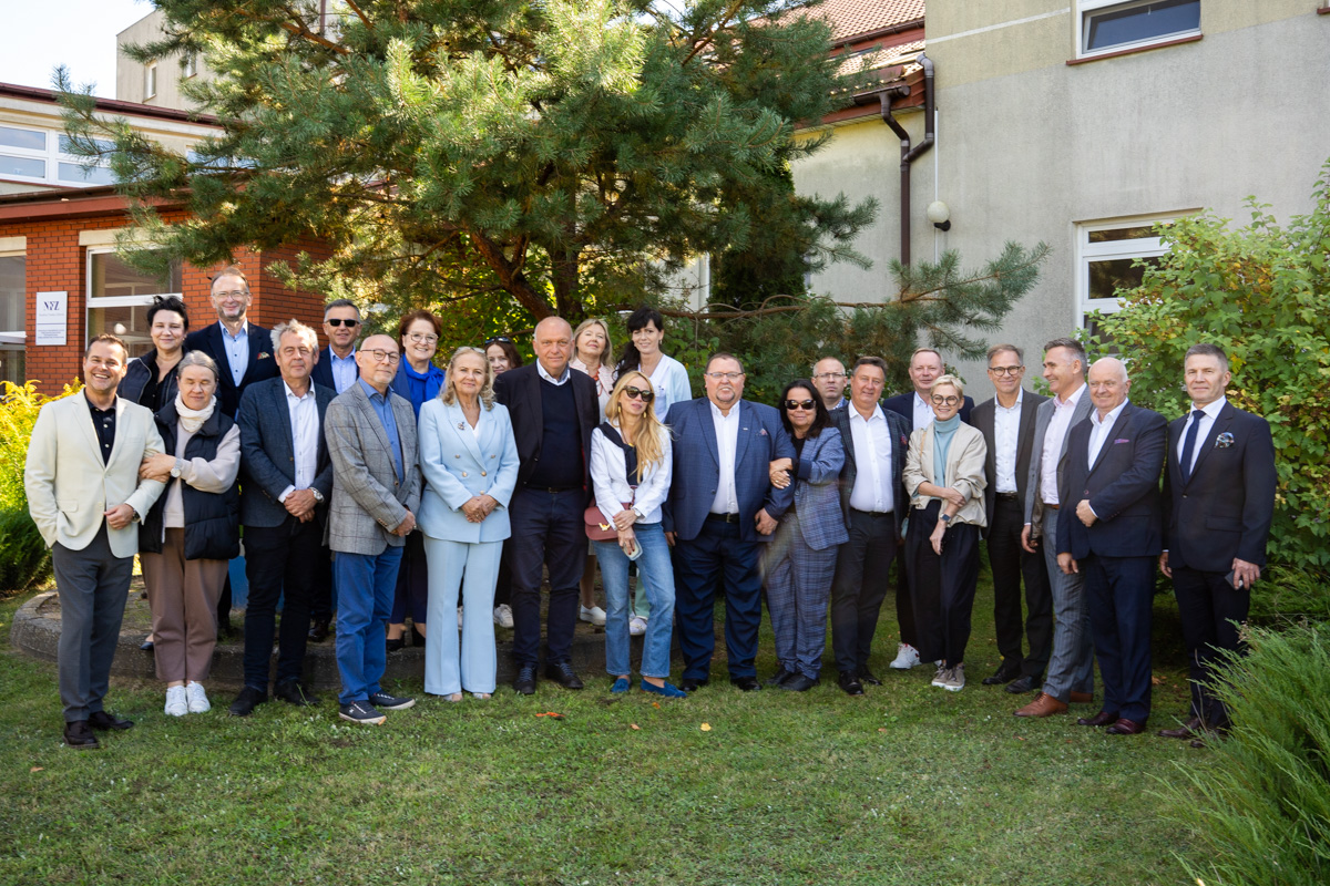 Grupa kobiet i mężczyzn stoi na trawie przed budynkiem, pozując do zdjęcia grupowego. W tle widoczne jest drzewo oraz fragmenty budynku z oknami. Osoby ubrane są zarówno w garnitury, jak i w stroje codzienne.