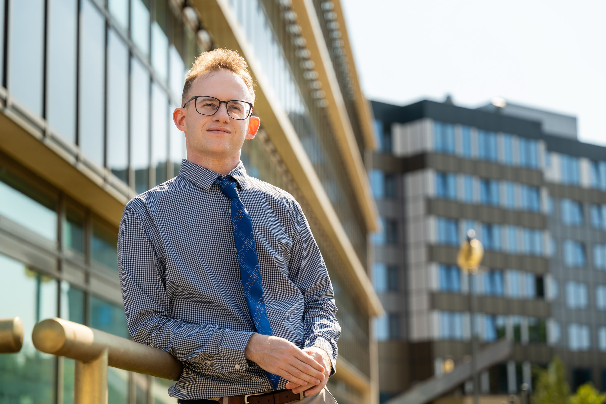 Piotr Alster, PhD hab. a young man wearing glasses, dressed in a shirt and tie, poses for a photo in front of buildings with glass windows. He smiles.
