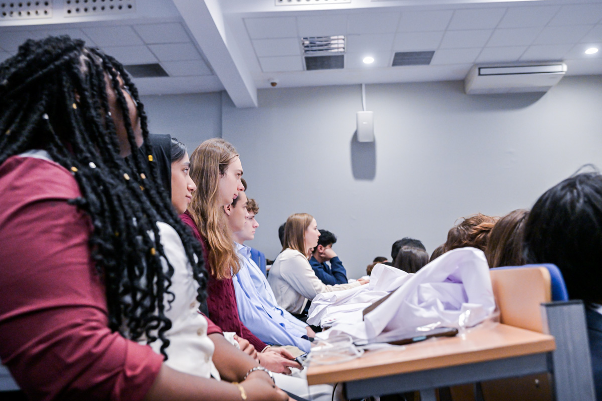 Grupa studentów w sali. Siedzą przy blatach. Na pierwszym planie kobieta z długimi, czarnymi włosami splecionymi w warkoczyki. 