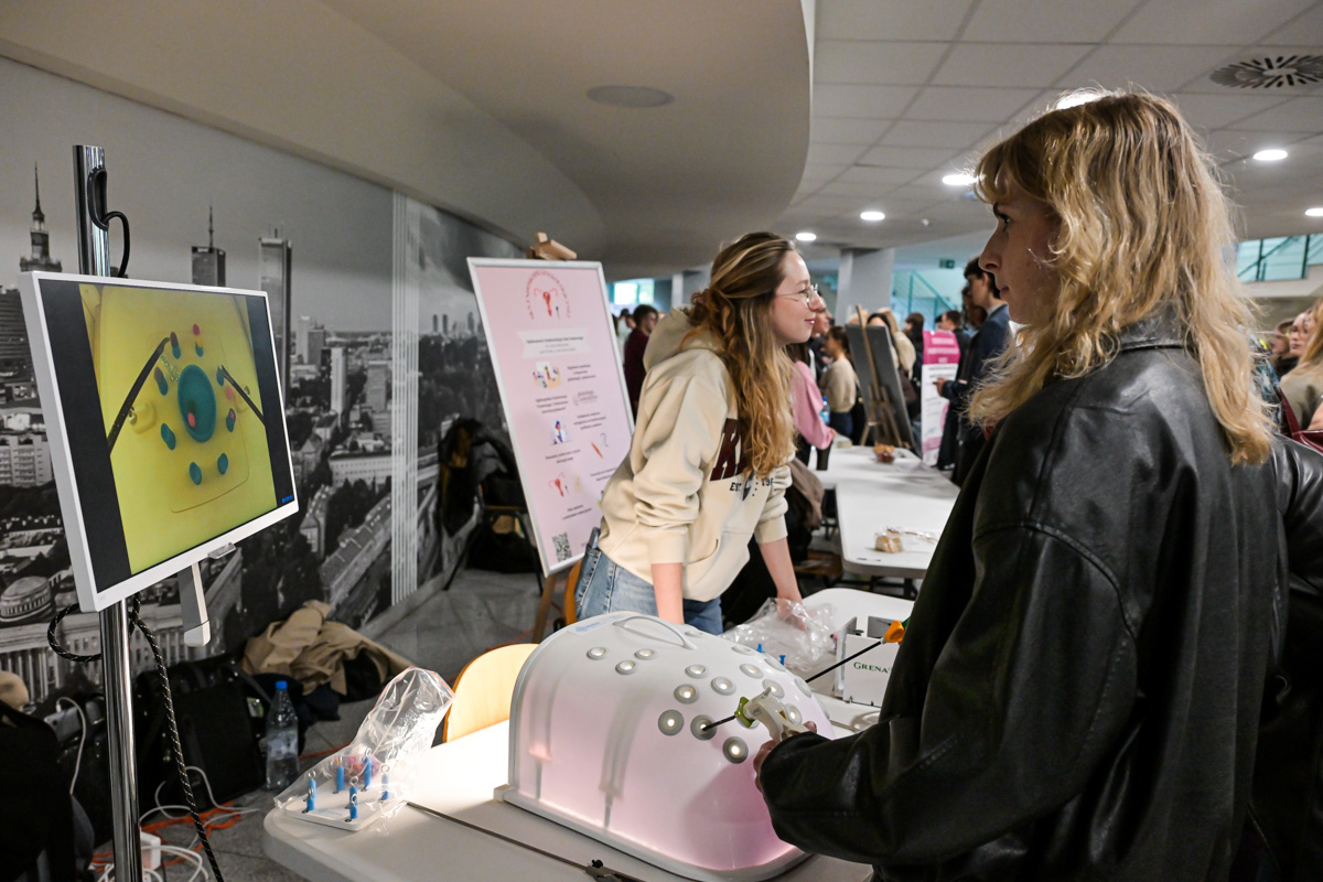 Two people are standing at a table with a white and pink medical simulator. One person in a light-colored sweatshirt is operating the device, while the other, wearing a black jacket, is looking at a screen displaying a color image of organs. In the background, there is a poster with instructions and a group of people at other stands.