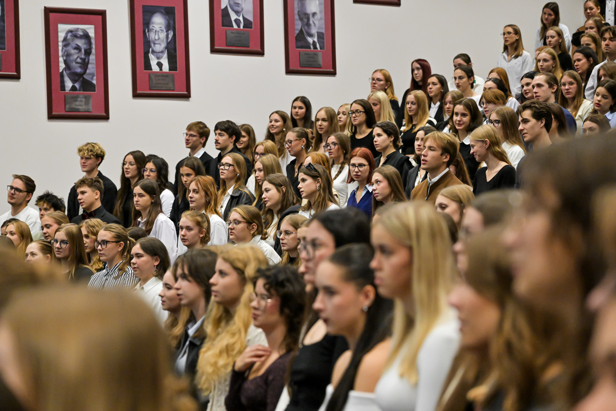 Studenci w czasie immatrykulacji. Około 30 młodych osób, są nieformalnie ubrani. Patrzą przed siebie, mają poważne wyrazy twarzy. W tle na ścianie widać portrety starszych mężczyzn w czerwonych ramkach.