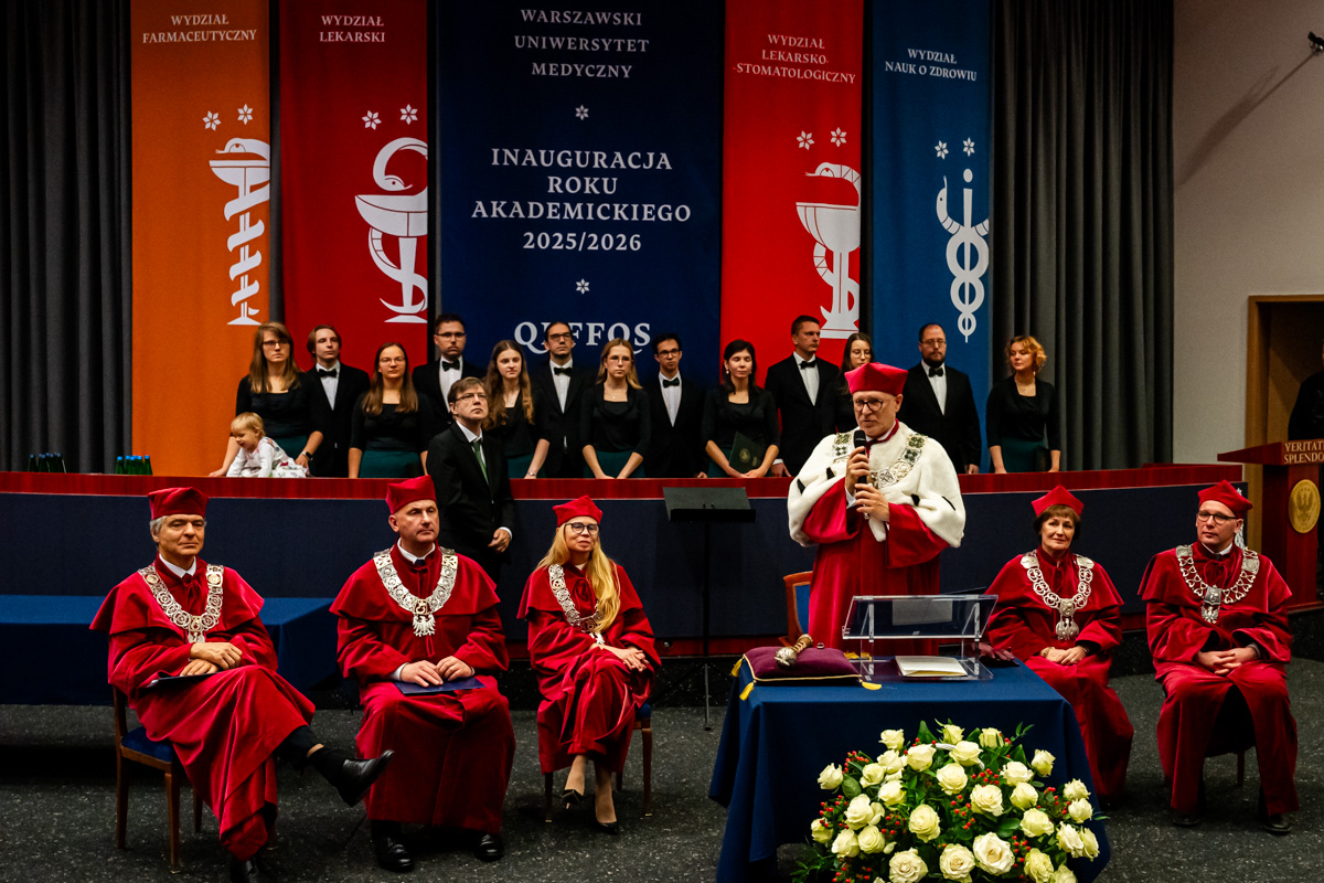 Sześć osób w czerwonych togach i biretach z insygniami rektorskimi na scenie auli. Jedna z nich, rektor, stoi i przemawia do mikrofonu. Pozostali siedzą. W tle widać chór w czarnych strojach oraz kolorowe banery z nazwami wydziałów. Na stole przed mównicą leży bukiet białych kwiatów. Scena przedstawia inaugurację roku akademickiego 2025/2026.