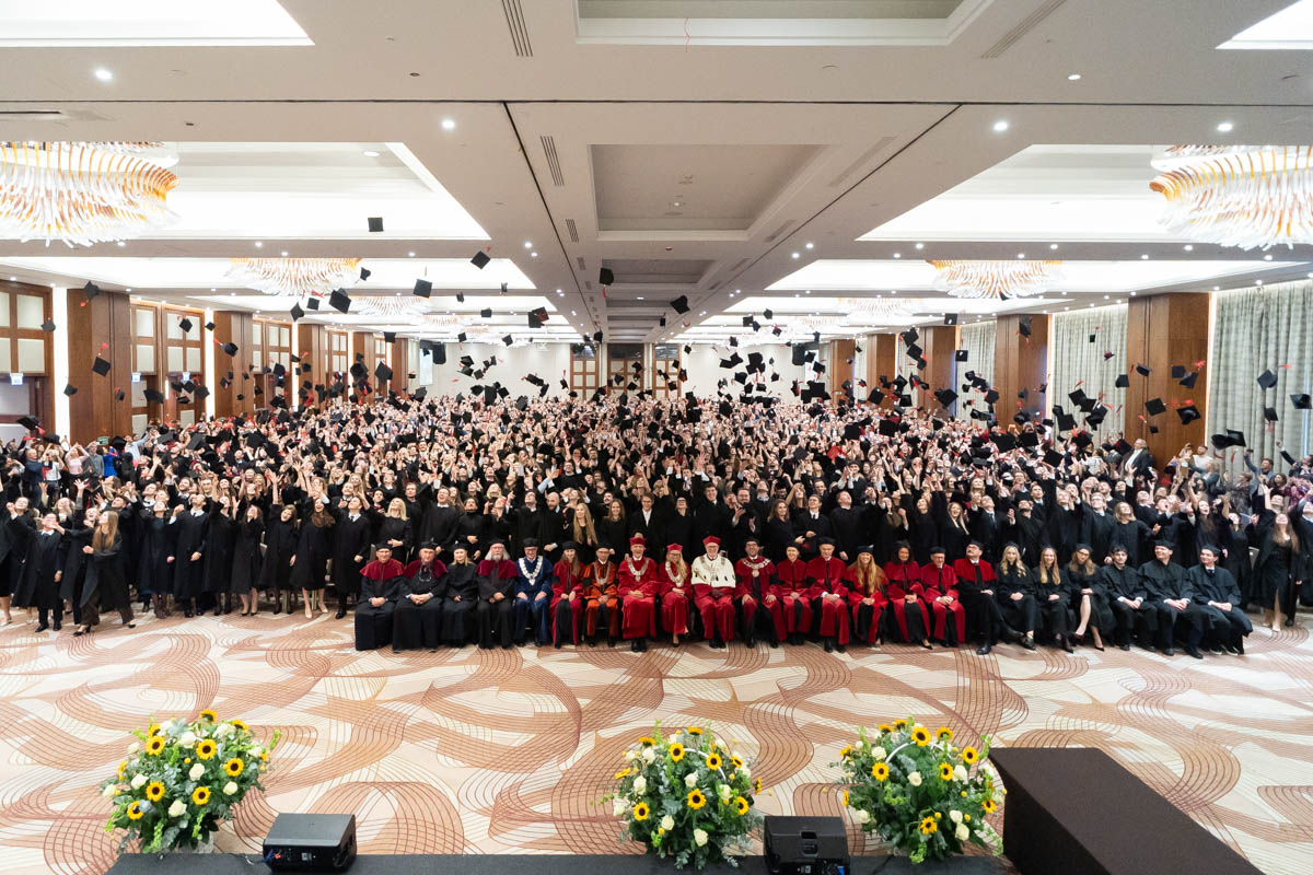 “A large group of people in black gowns stands in a conference hall and throws graduation caps into the air. In the first row, several people are seated wearing red and navy gowns. In the foreground, there are two floral arrangements with yellow sunflowers.