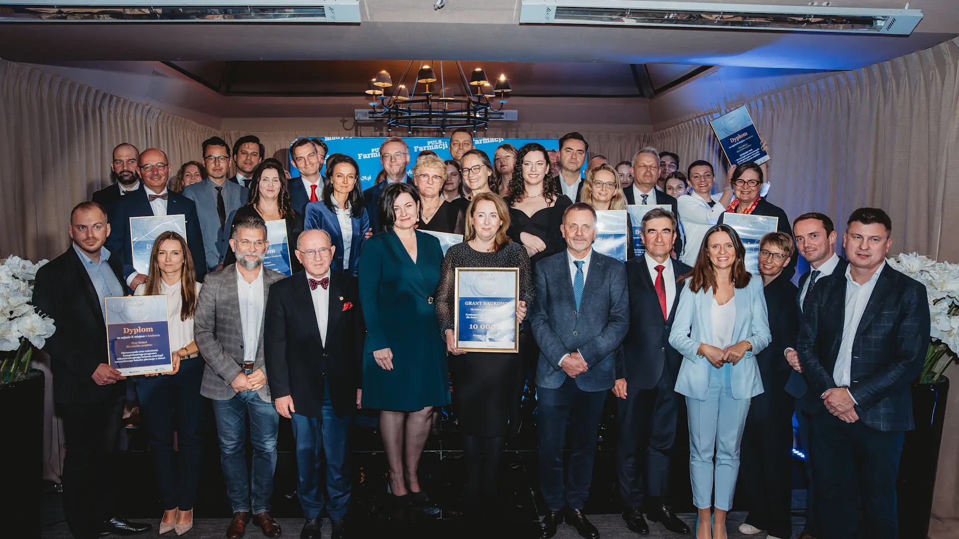 A group of elegantly dressed people pose for a group photo. Some of them are holding framed diplomas in their hands.