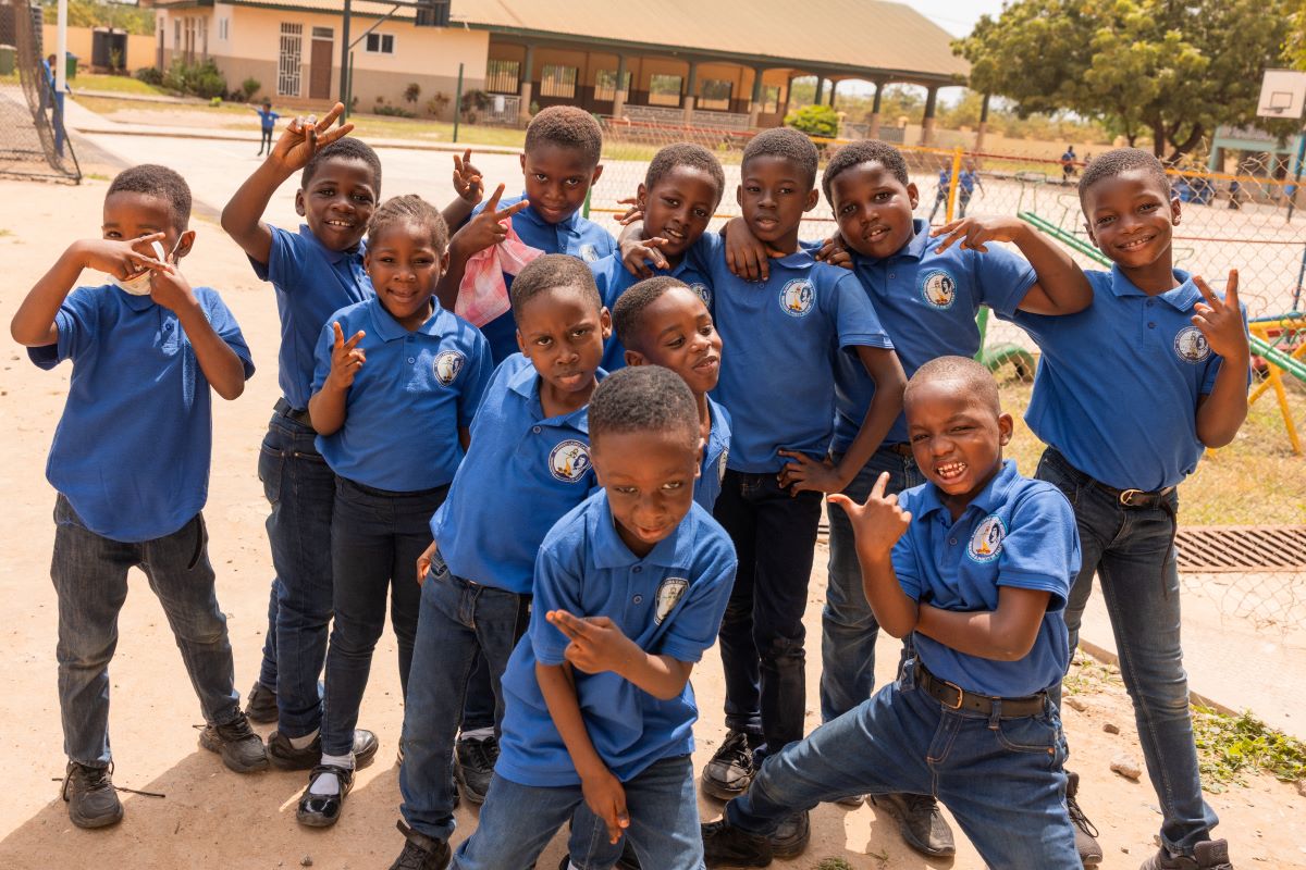 A group of dark-skinned children in blue school uniforms is standing outside, posing with various dynamic gestures against the backdrop of a school building and a playground.