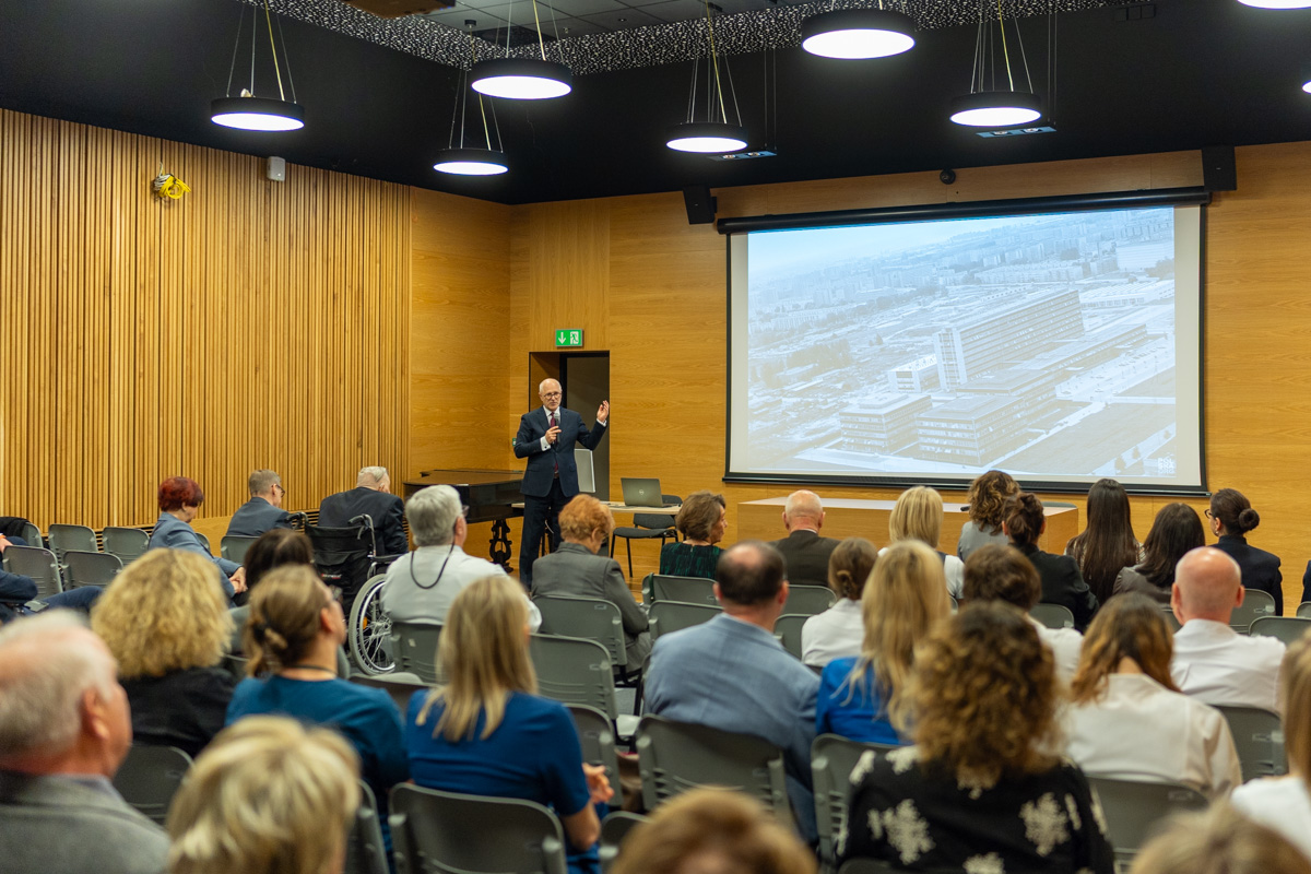A large hall where several dozen people are gathered. Everyone is seated except for Professor Rafał Krenke, who is standing in the middle of the room and speaking into a microphone. Behind him, a large multimedia screen is visible.