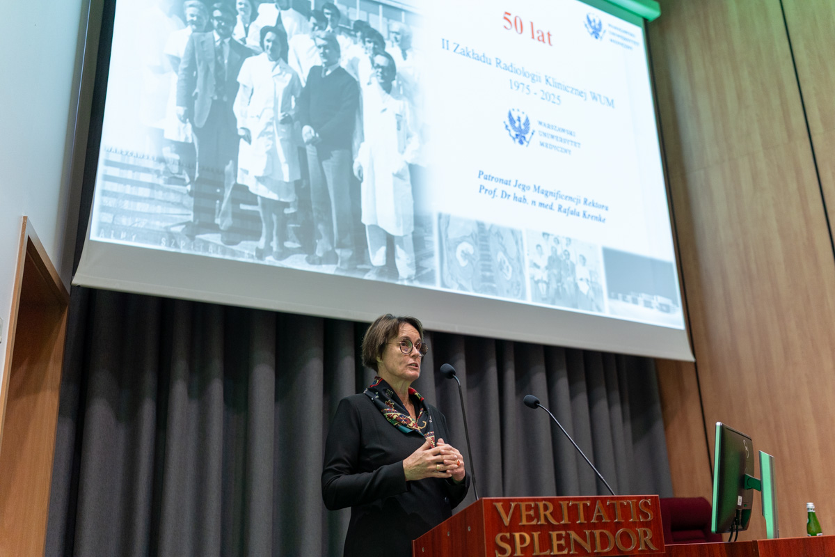 A woman, Prof. Magdalena Januszewicz, is standing behind a lectern and speaking. In the background, a multimedia screen displays a presentation.