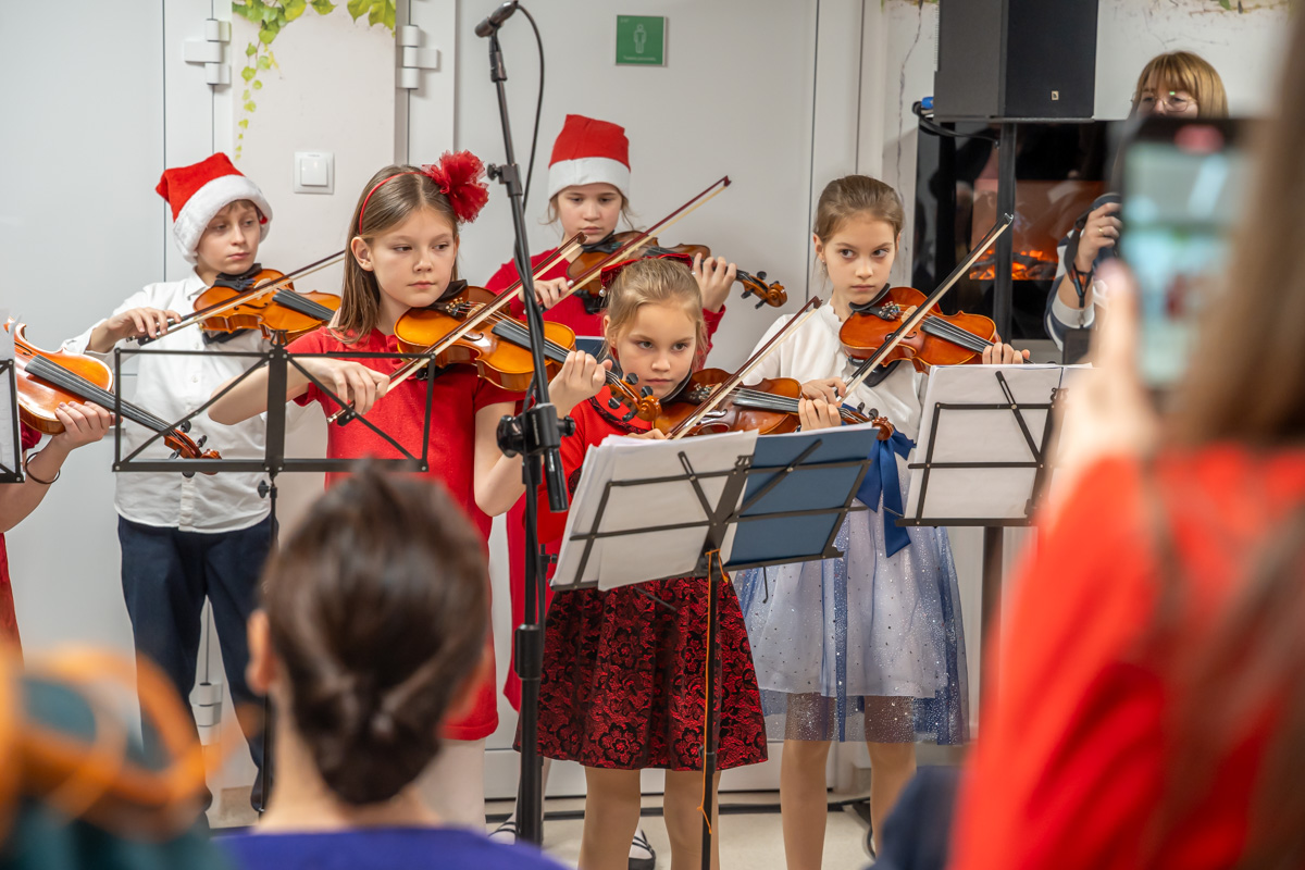 A group of children is playing the violin during a Christmas concert. They are standing in a row in front of music stands, some wearing red Santa hats. In the background, a microphone and a speaker are visible, and in front of the stage, the audience is seated.