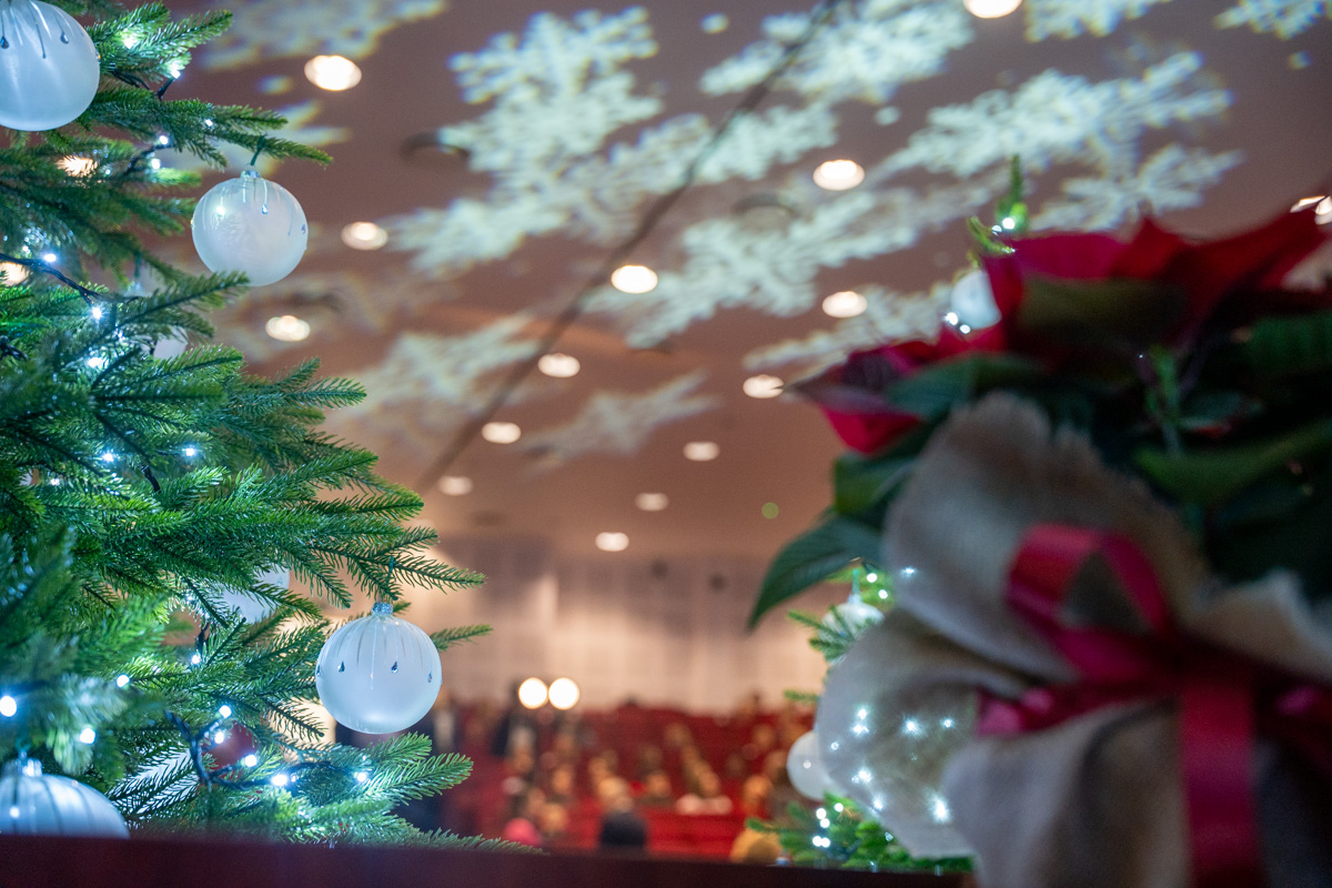 A Christmas tree with white baubles stands in the foreground. Next to it is a bouquet of red flowers wrapped in jute and tied with a red ribbon. In the background, there is a hall with red chairs and blurred silhouettes of people. Bright snowflake-shaped projections are shining on the ceiling.
