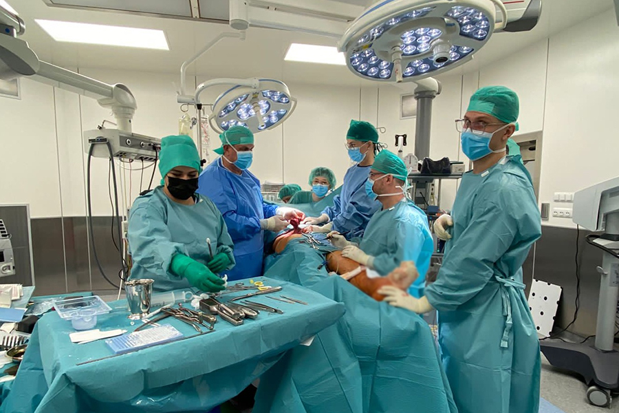 Prof. Krakowczyk z zespołem w czasie procedury. Osoby są ubrane w niebieskie fartuchy lekarskie, część z nich jest pochylona nad pacjentem, który leży na stole operacyjnym. Obok lekarzy widać stół z narzędziami, a także różne sprzęty medyczne.