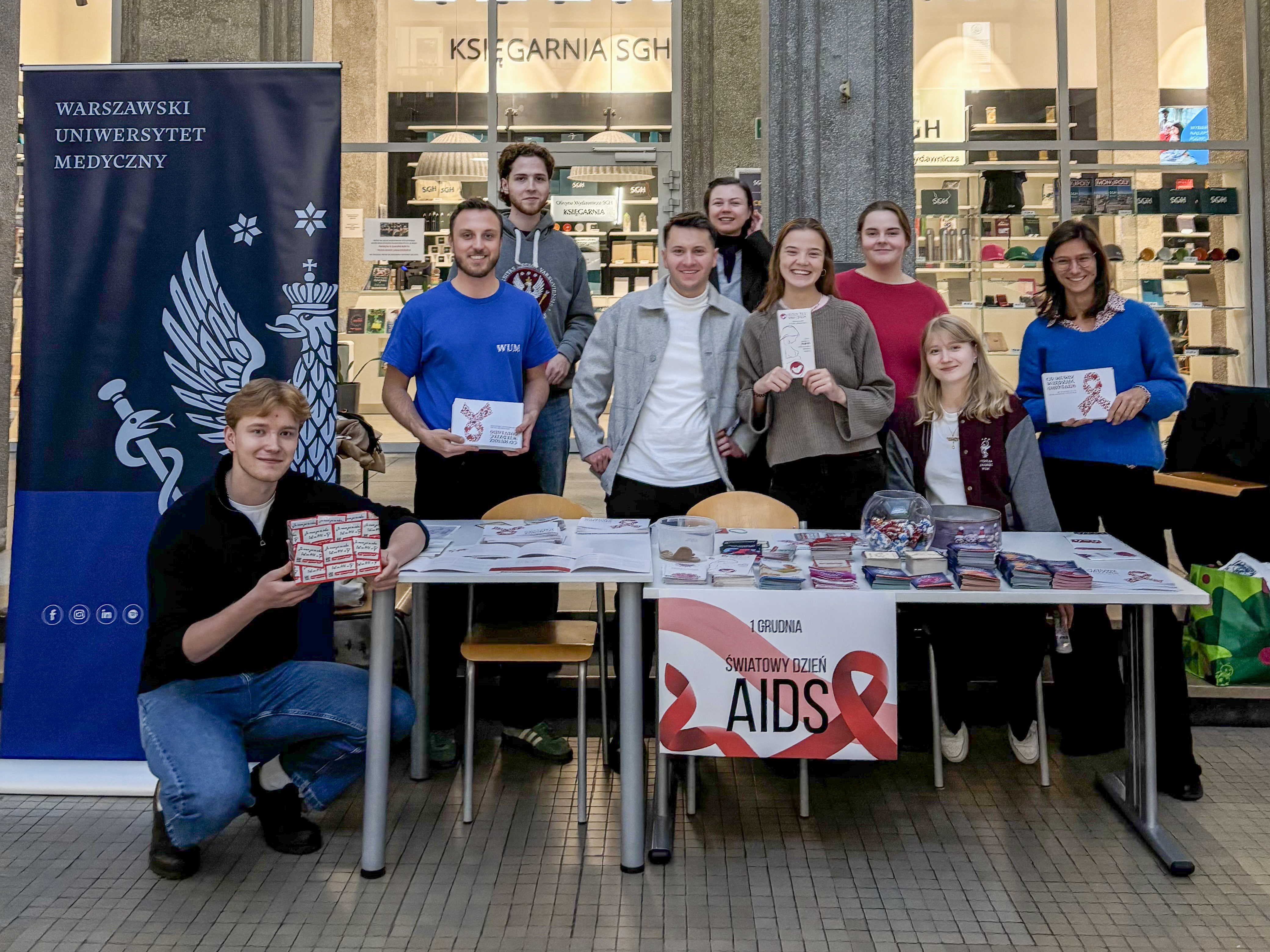 Grupa studentów stojących przy stoisku informacyjnym z ulotkami, materiałami edukacyjnymi i gadżetami promującymi zdrowie w przestrzeni uczelni.