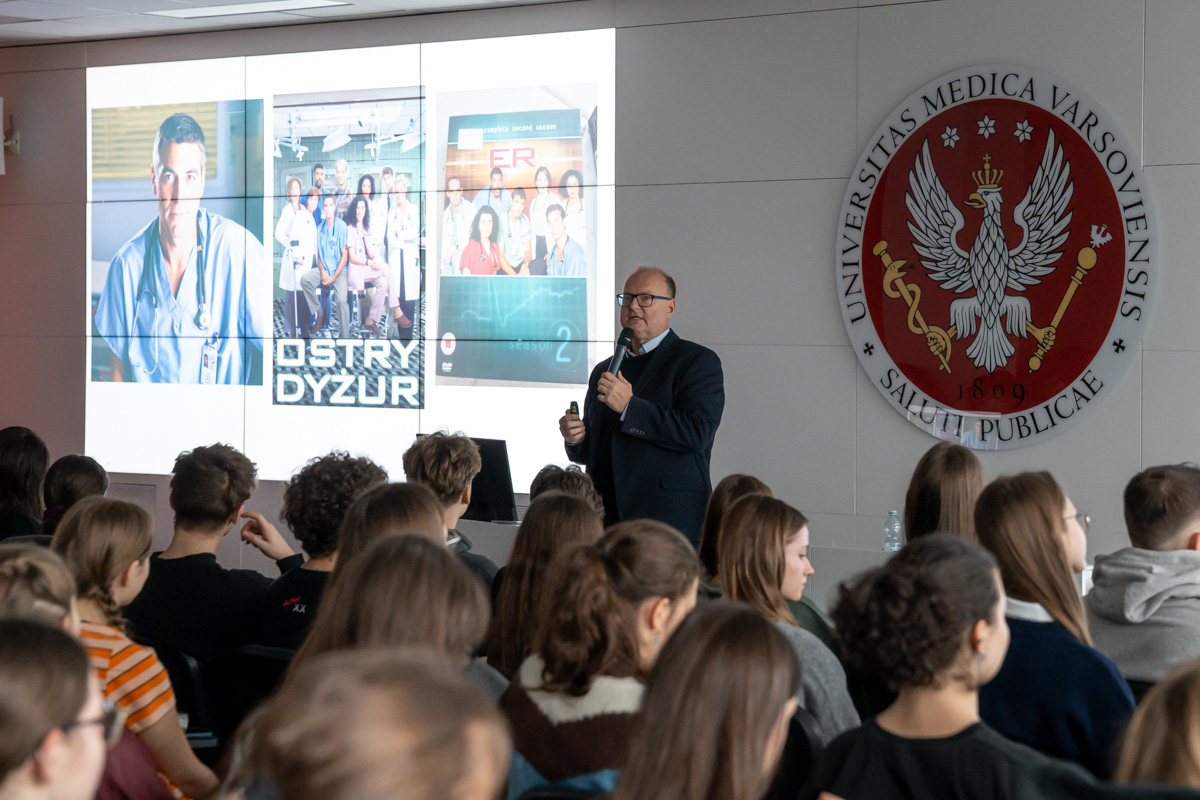 Prof. Czupryniak w czasie wykładu stoi na środku sali. Za nim widać czerwone godło WUM oraz ekran multimedialny z wyświetlonymi zdjęciami. Przed nim w rzędach siedzą młodzi ludzie.