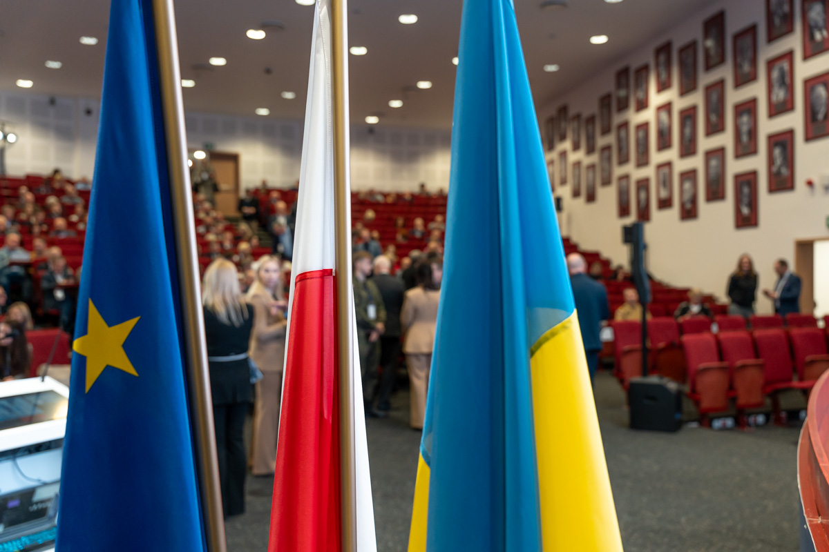 Three flags, from left: the European Union, Poland, and Ukraine, stand in a large hall. People gathering can be seen in the background.