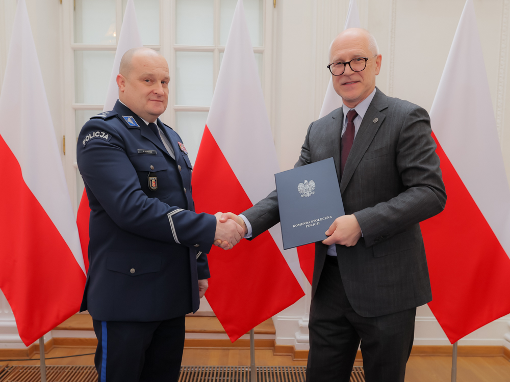Two men, the one on the left wearing a police uniform, the one on the right wearing a suit, are shaking hands. The man on the right is holding a closed briefcase. Polish flags can be seen in the background.
