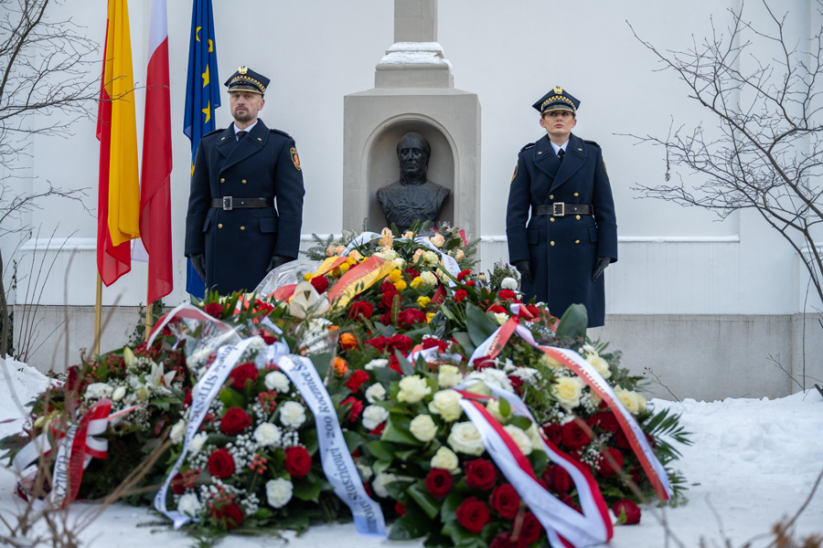 Dwa umundurowane osoby stoją po obu stronach pomnika z popiersiem. Przed nimi leży duży, rozsypany półkolem stos wieńców i bukietów w barwach białych, czerwonych, żółtych i zielonych, z widocznymi szarfami. Obok pomnika stoją maszty z flagami. 
