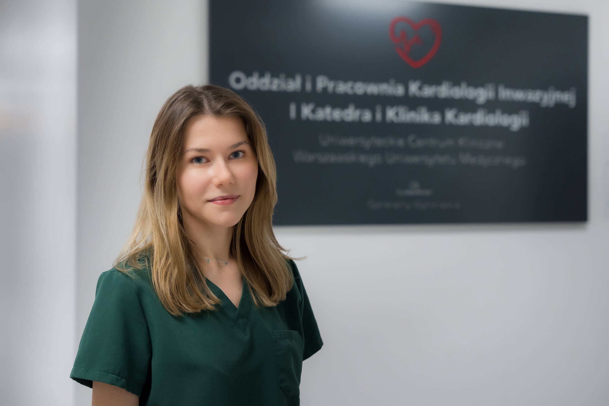 Lek. Ewelina Błażejowska, ubrana w zielony fartuch lekarski, pozuje do zdjęcia, patrzy prosto w obiektyw. Za nią na ścianie widać tablicę informacyjną z czerwonym sercem i białym napisem.