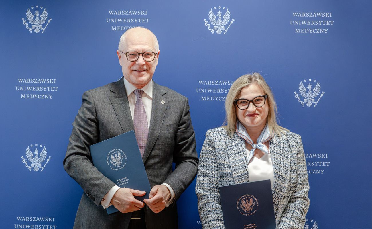 Mężczyzna - rektor WUM oraz kobieta - zastępczyni dyrektora ds. naukowych NIGRiR, elegancko ubrani, pozują do zdjęcia na tle niebieskiej ścianki z białym logiem WUM i napisem "Warszawski Uniwersytet Medyczny".