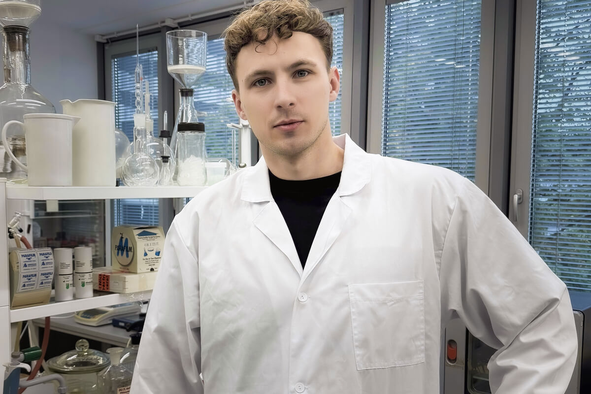 Osoba w białym fartuchu laboratoryjnym stojąca w nowoczesnym laboratorium chemicznym, otoczona kolbami, cylindrami miarowymi i innym sprzętem badawczym, na tle półek i dużych okien.