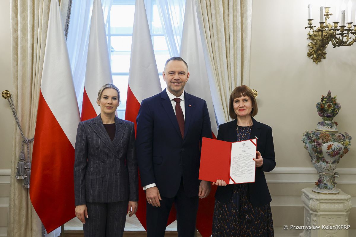 Three people in the photo are standing in an ornate hall. In the center is the President of the Republic of Poland, to the left the President’s spouse, and to the right – Prof. Aleksandra Wesołowska. On the left is the Polish flag. Prof. Aleksandra Wesołowska is holding an open folder with the appointment document.