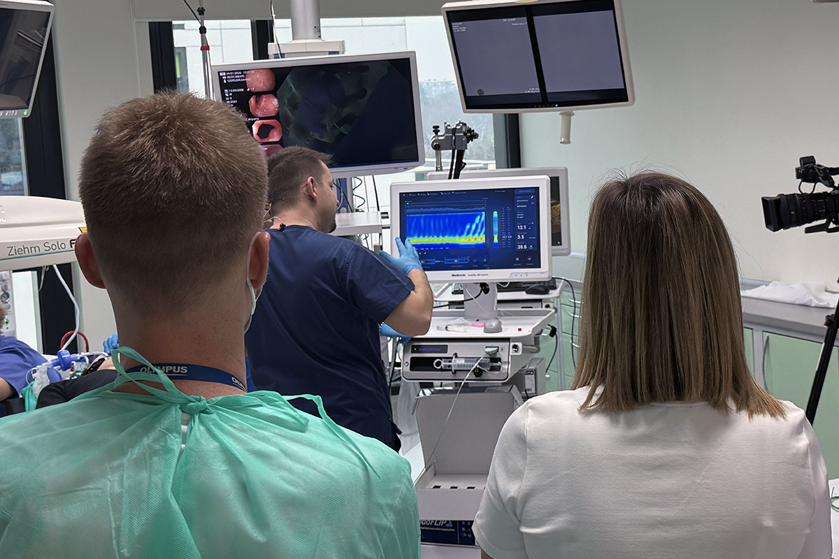 Kilka osób ubranych w fartuchy medyczne w sali zabiegowej. Wokół widać sprzęt medyczny.