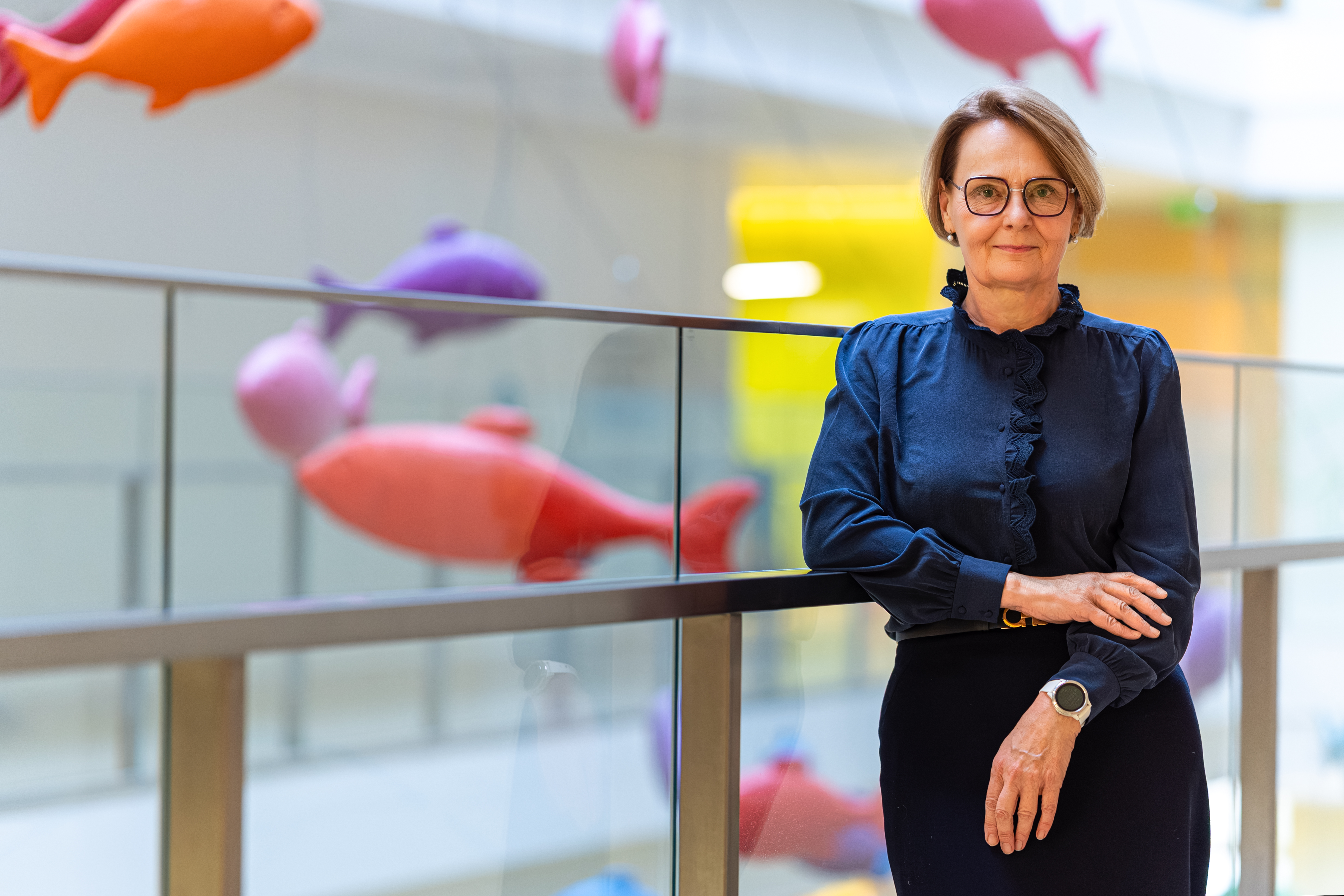 A person standing by a glass railing in a brightly lit indoor space decorated with large, colorful fish sculptures suspended in the background.