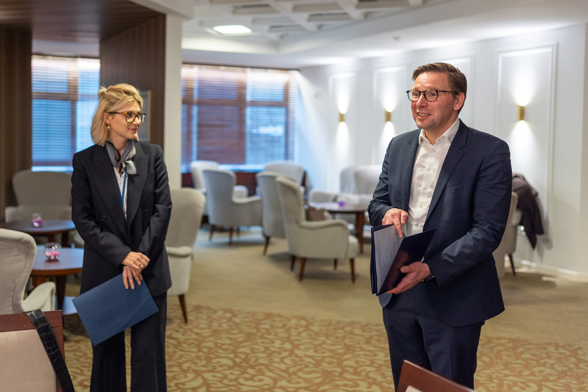 Dwie elegancko ubrane osoby: po lewej kobieta, po prawej mężczyzna, stoją w sali z jasnymi ścianami i złotym dywanem.