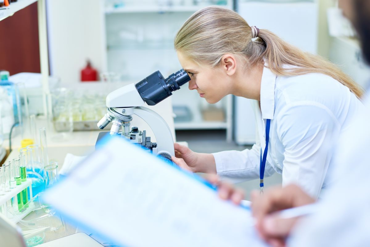 Osoba w laboratorium obserwuje próbki pod mikroskopem, w otoczeniu probówek i sprzętu laboratoryjnego