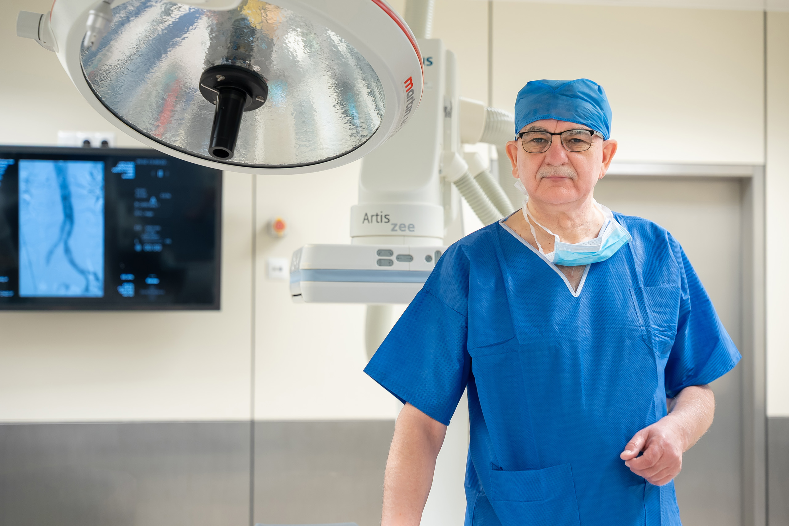 A medical professional wearing blue surgical scrubs and a cap stands in an operating room equipped with overhead surgical lighting and advanced imaging equipment